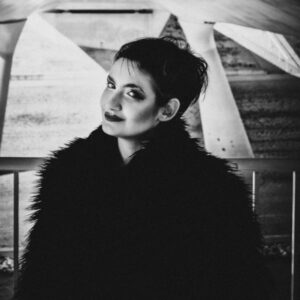 Sanjina Kashikar steht mit kurzen Haaren und Pelzmantel vor einer Industriekulisse. Bild: © Patrik Zedlewski