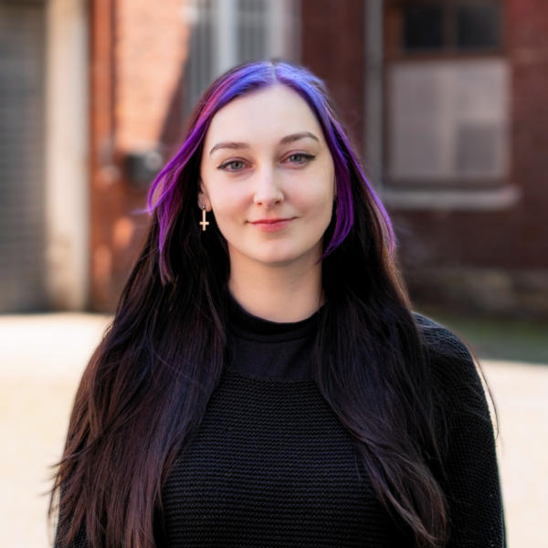 Mirjam Kay Mashkour mit violetten Haaren vor einer Backsteinmauer.
Bild: © Thiemo Rudolph