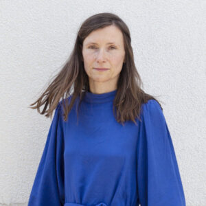 Portraitfoto der Autorin Heike Geißler in blauem Kleid und wehenden Haare vor einer weißen Hauswand. Bild: © Cihan Çakmak