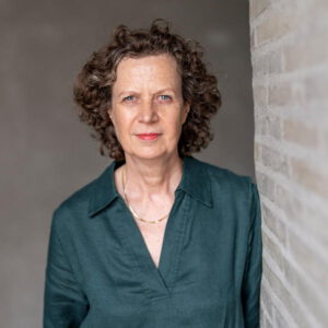 Die Historikerin Dr. Maren Gottschalk mit lockigem Haar und grüner Blouse an eine Ziegelmauer gelehnt. Bild: © Peter Susewind