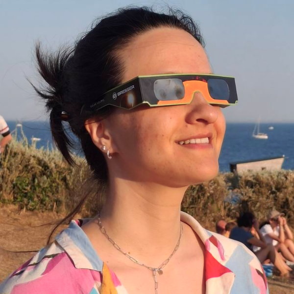 Die Autorin Alexandra Begau im Freien am Meer mit bunter Bluse und Sonnenfinsternisbrille blickt lächelnd in den Himmel.
Bild:  © privat